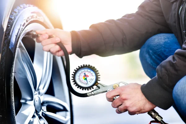 A man kneeling next to a car wheel checks the tire's air pressure with a hose-connected pressure gauge.