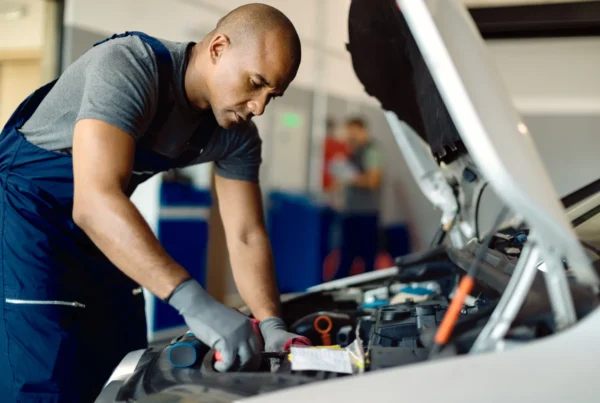 A mechanic in navy coveralls and gray gloves leans under the open hood of a white car working on the engine in an auto repair shop.
