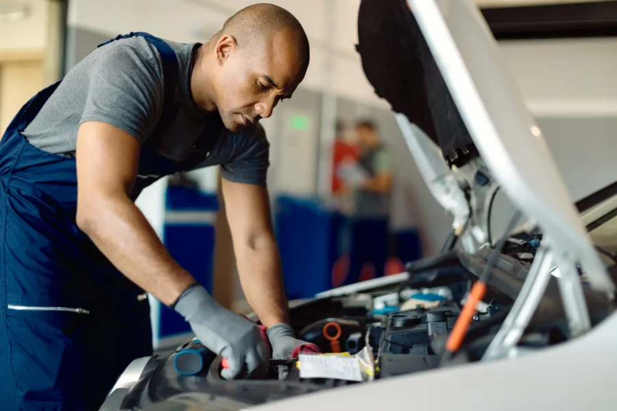 A mechanic in navy coveralls and gray gloves leans under the open hood of a white car working on the engine in an auto repair shop.