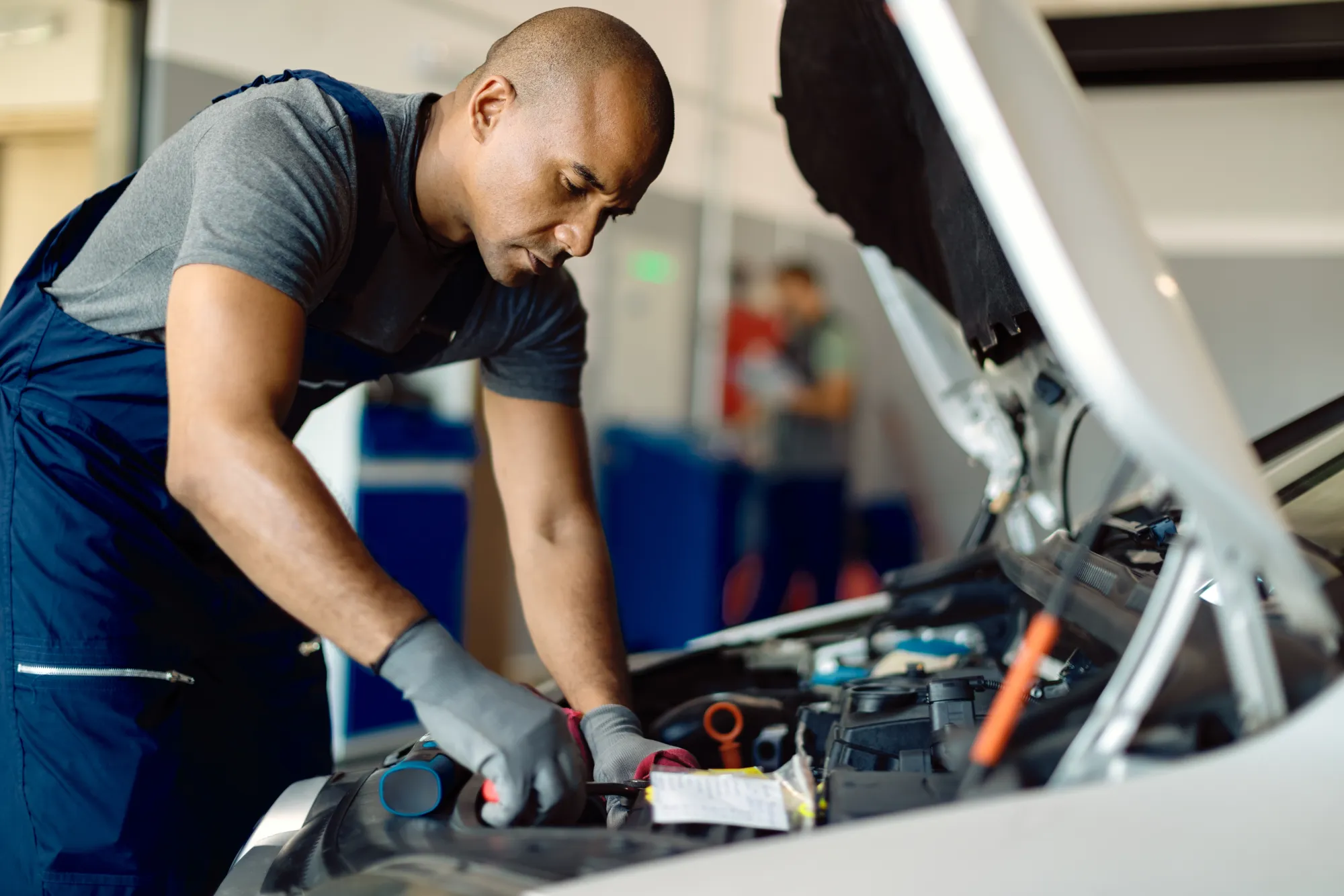 A mechanic in navy coveralls and gray gloves leans under the open hood of a white car working on the engine in an auto repair shop.