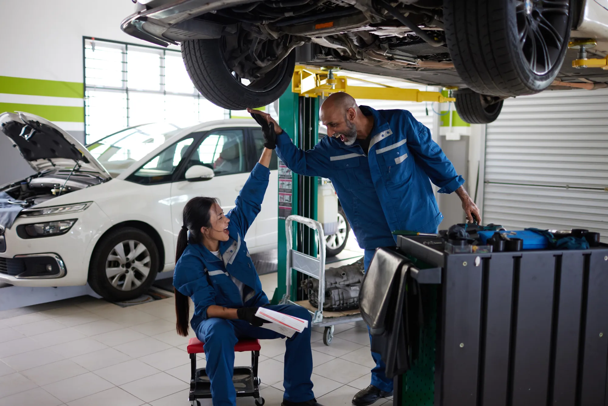 Two mechanics in blue coveralls high-five under a car raised on a lift in an auto repair shop.