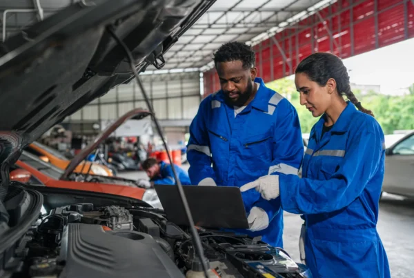 Two mechanics in blue coveralls use a laptop to run diagnostics on a car engine with the hood open in a repair shop.