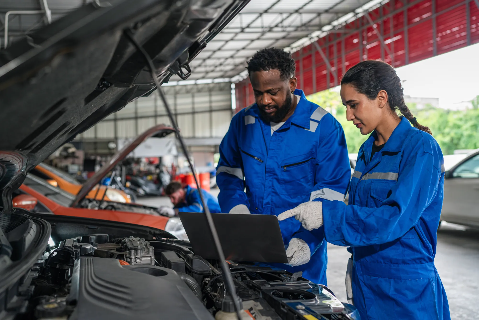 Two mechanics in blue coveralls use a laptop to run diagnostics on a car engine with the hood open in a repair shop.